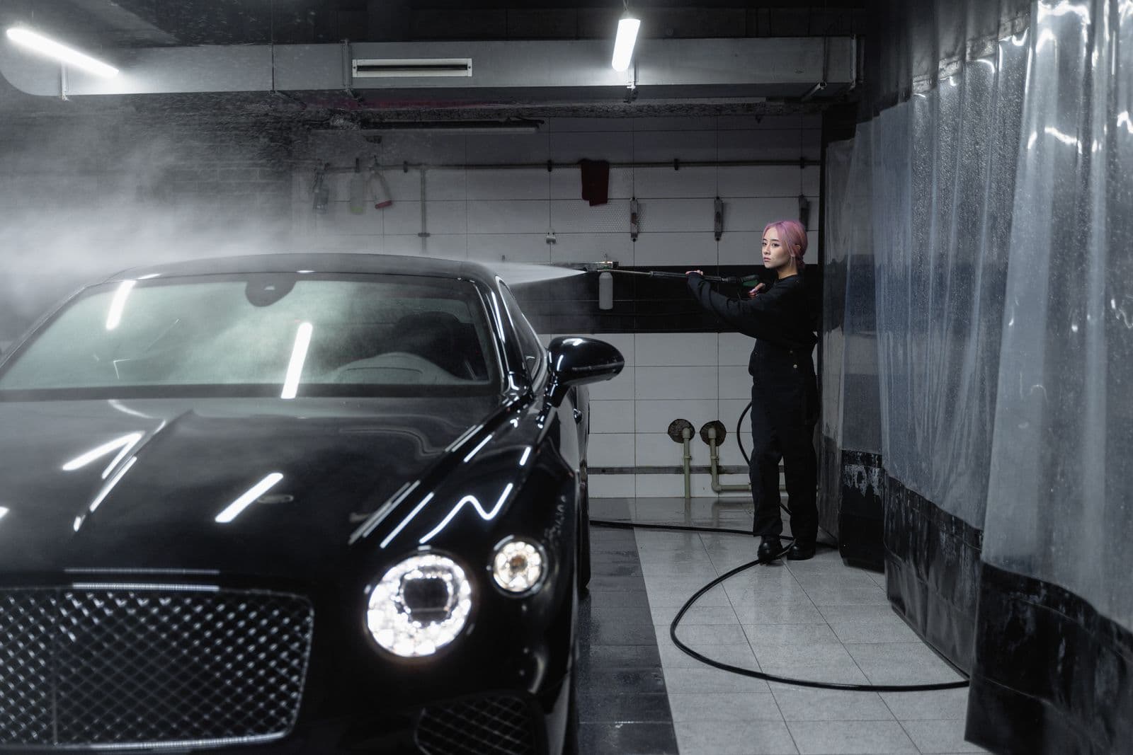 Detailer rinsing a black coupe with a pressure washer in a wash bay