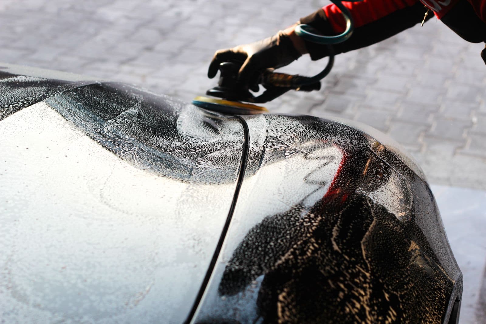 Detailer working a polisher over a dark car outdoors on the pavement
